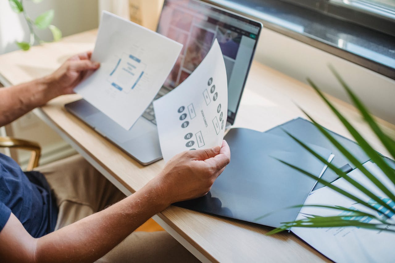 creative Crop anonymous male designer in casual wear sitting at table with opened laptop and folders and reading papers